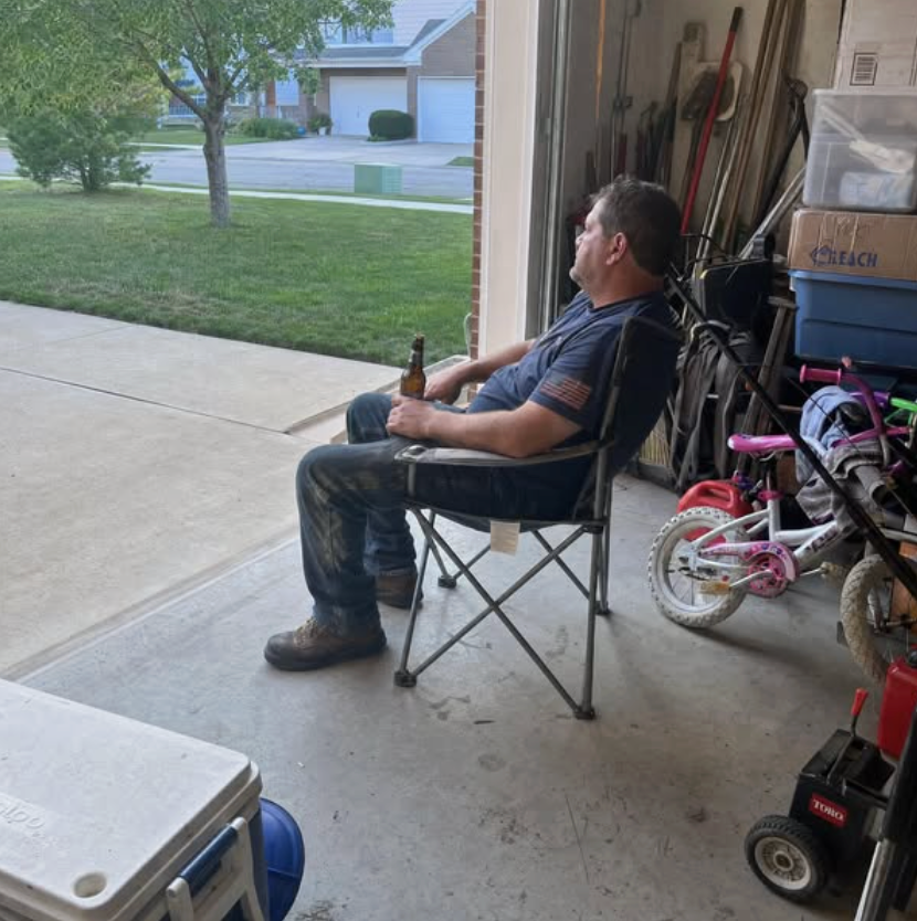 A man sitting alone in a folding chair in his open garage at the end of the day, holding a beer, looking out at the empty street.
