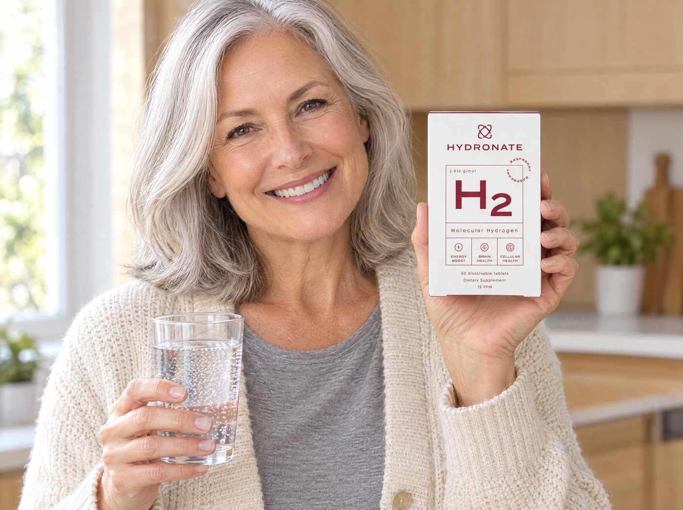 A smiling woman in her sixties holding a box of Hydronate hydrogen tablets and a glass of sparkling water in her kitchen.