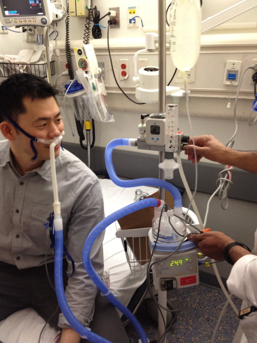 A patient sitting on a hospital bed receiving hydrogen inhalation therapy through a nasal cannula, with clinicians adjusting a hospital-grade delivery device beside him.
