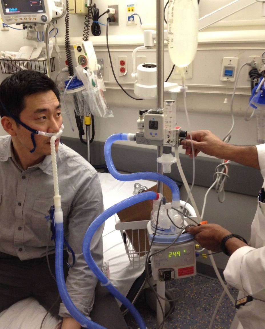 A patient sitting on a hospital bed receiving hydrogen inhalation therapy through a nasal cannula while a clinician adjusts the medical-grade delivery unit.